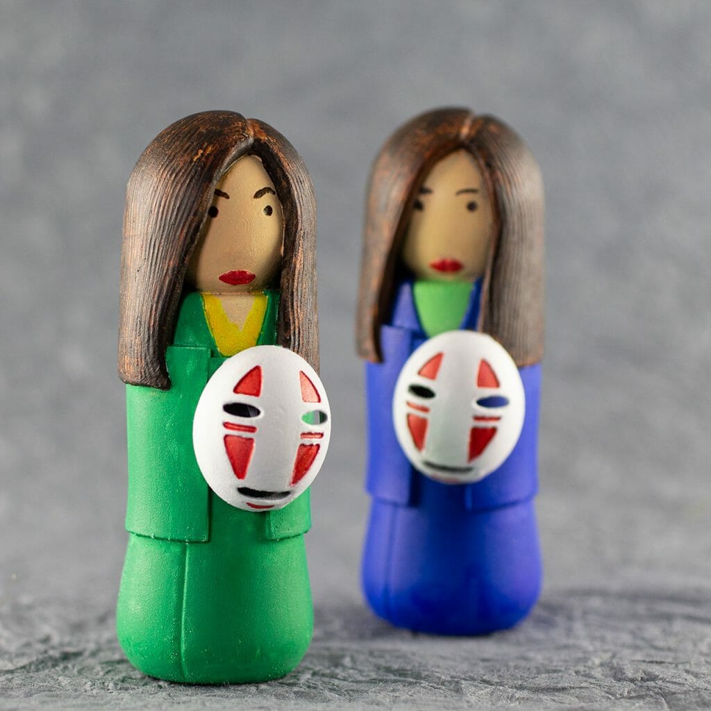 Two small kokeshi (a traditional style of Japanese doll), painted by hand. One has a bright green kimono, the other is blue. Simple facial features. Long brown hair. They are each holding a No Face mask from the animated film Spirited Away.