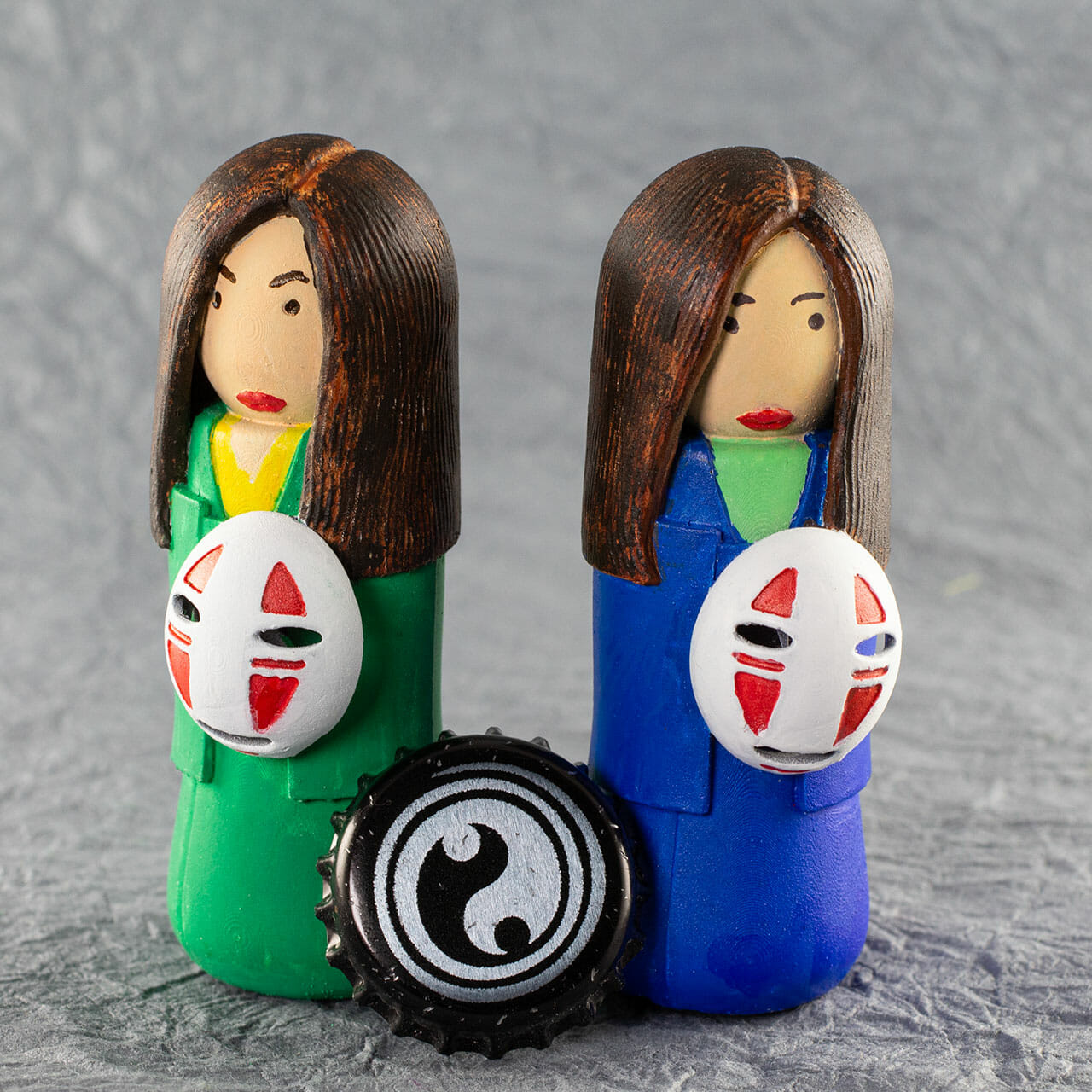 Two small kokeshi (a traditional style of Japanese doll), painted by hand. One has a bright green kimono, the other is blue. Simple facial features. Long brown hair. They are each holding a No Face mask from the animated film Spirited Away. Bottle cap for scale.