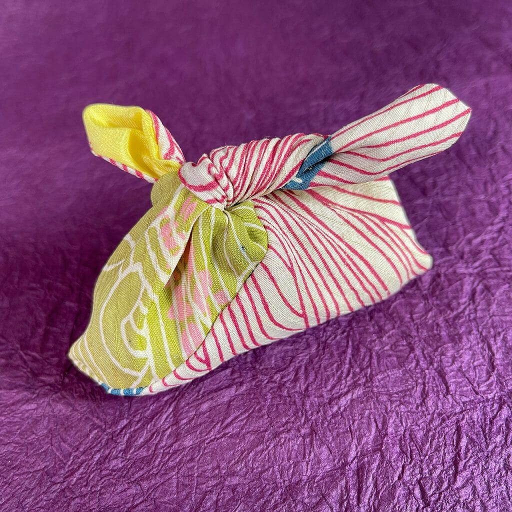 A tiny traditional bag that ties shut, made from a bit of silk kimono fabric with a white base printed with red lines, green swirls and little bits of pink and blue, lined with solid yellow silk.