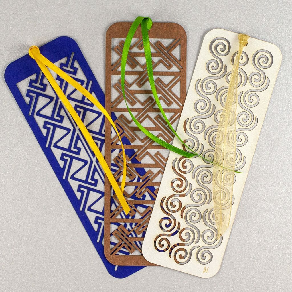 Collection of three Celtic kirigami bookmarks: two different key pattern designs in cobalt blue and copper brown and a pattern of spirals in a white pearlescent stock.