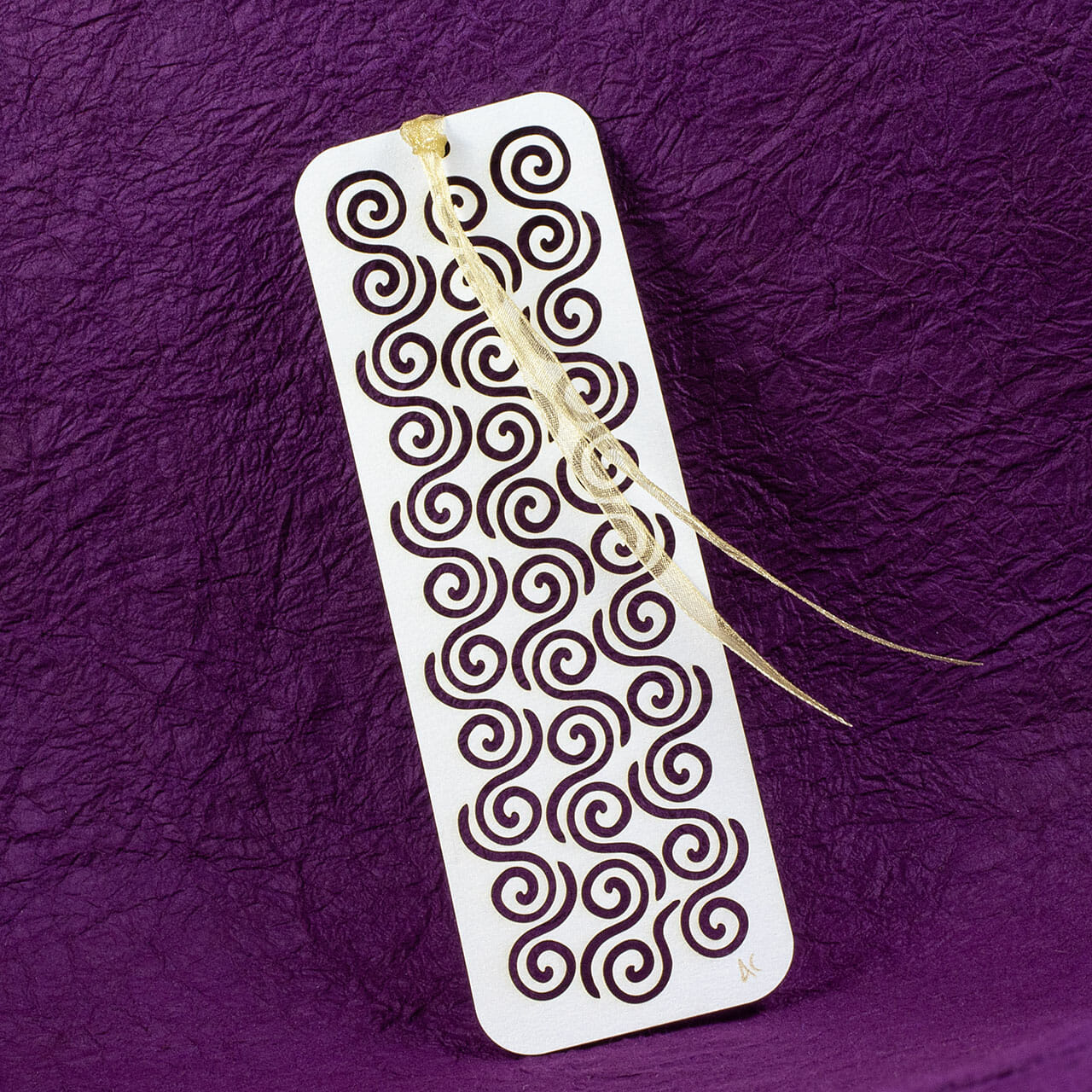 Kirigrami bookmark with an interlocking pattern of organic Celtic spirals cut from a white pearlescent stock with a gold gauze ribbon tied to one end.