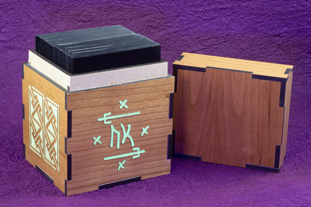 Wood sleeved "commander" deck box for M:tG and similar games with glowing engraved designs: an embellished Futhark rune on the front and, a Celtic key pattern panel on the left side. Lid removed, showing a sleeved deck inside.