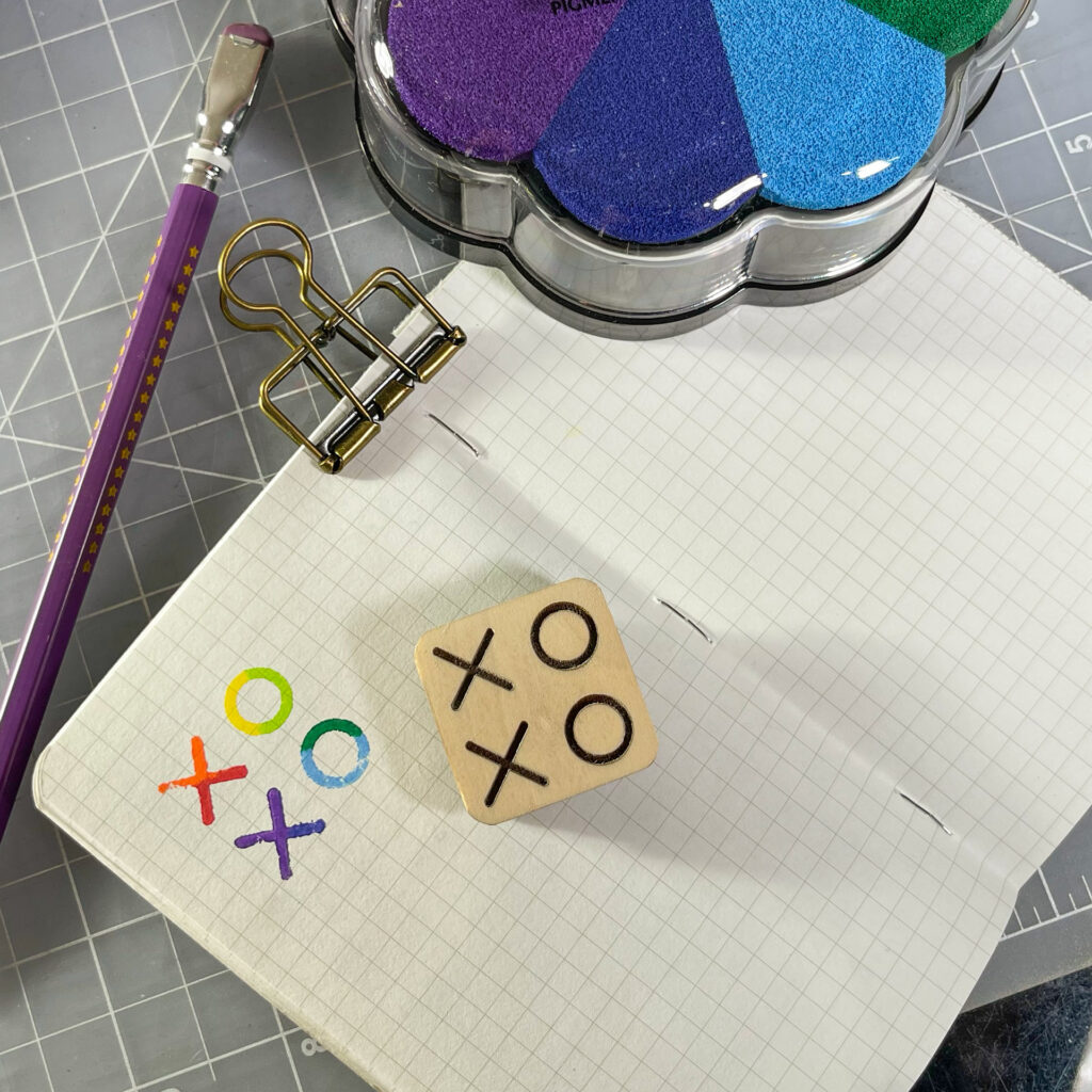 A small notebook lying clipped open stamped with XOXO in rainbow colors. The stamp sits next to the impression. A multi-colored stamp pad and a purple pencil are visible around it.