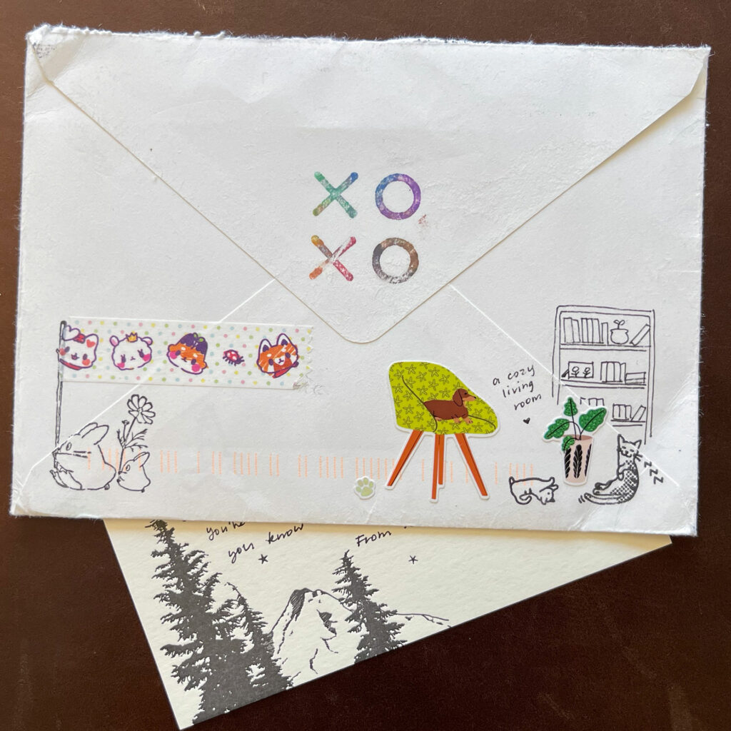 The back of an envelope with XOXO stamped in multiple colors on the flap, decorated with stickers. The corner of a card sticks out from the bottom with a stylized mountain and some pine trees.