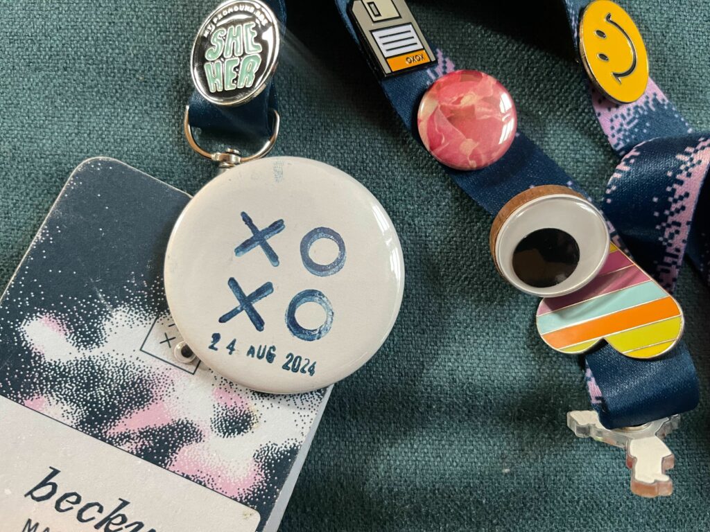 Stamped button, blue on white with the date 24 AUG 2024 stamped below, sitting on top of an XOXO festival pass and lanyard adorned with assorted pins.