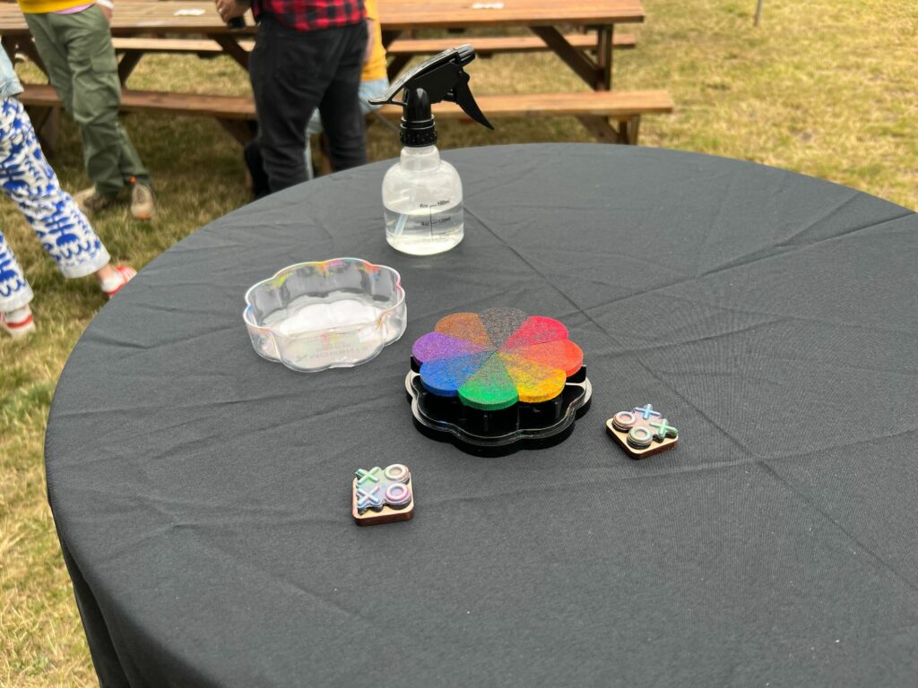 XOXO stamps, face-up, with flower-shaped rainbow stamp pad and spray bottle on a round table with a grey tablecloth in the park