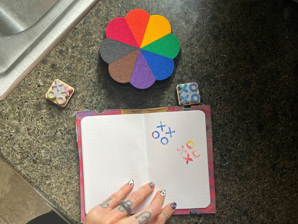 XOXO stamps with impressions in a small grid-ruled notebook on a granite surface with a rainbow flower stamp pad and fingers with intricately-patterned nails.