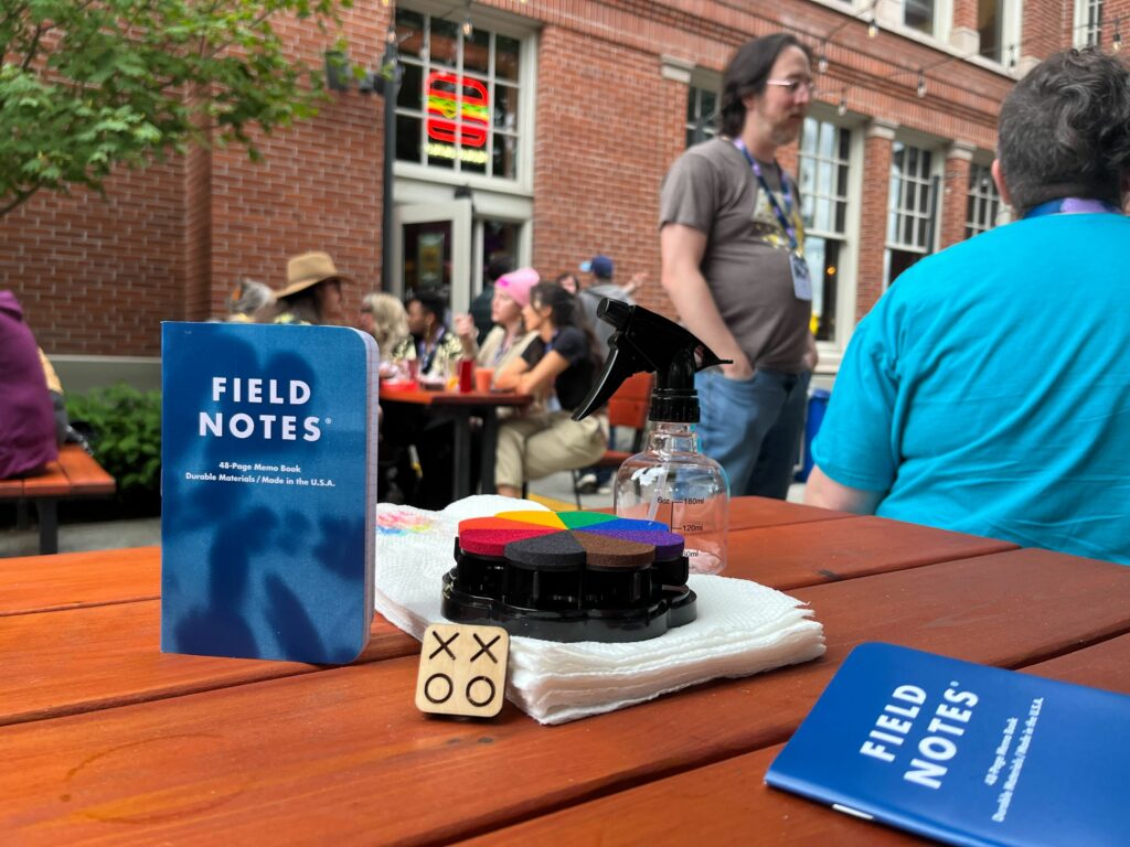 XOXO stamp, rainbow stamp pad, spray bottle, paper towels and, 2024 edition XOXO Field Notes on a picnic table.