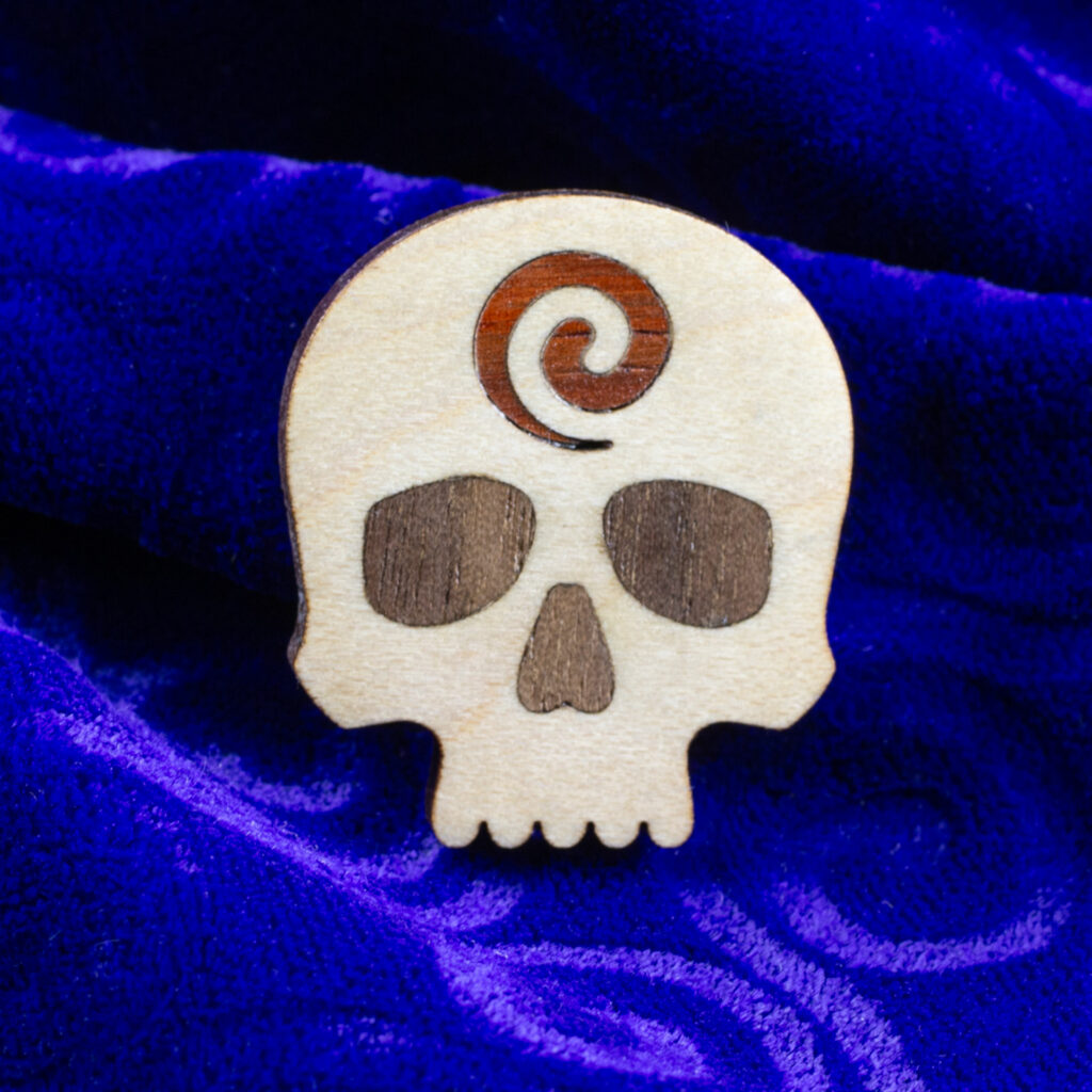 Godar Blot skull pin – white oak skull with inlaid walnut eyes and nose with a padauk spiral on the forehead.