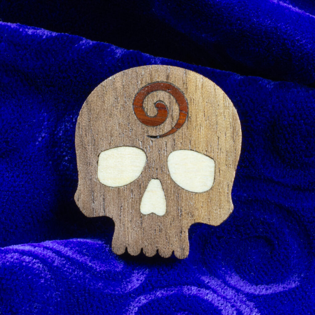 Godar Blot skull pin – walnut skull with inlaid white oak eyes and nose with a padauk spiral on the forehead.