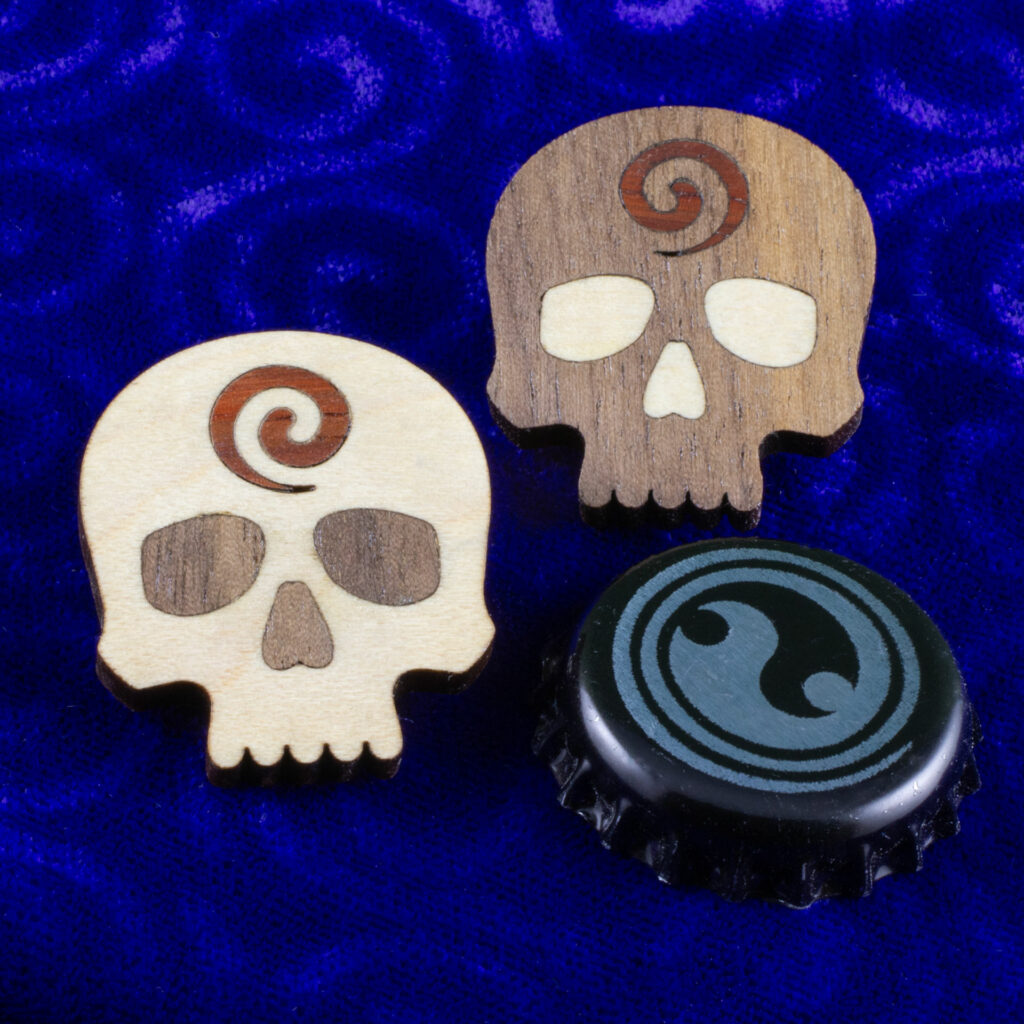 Godar Blot skull pins – hardwood inlay skull pins from white oak, walnut and, padauk. The white oak is dominant on one pin and the walnut is dominant on the other. A bottle cap slightly smaller than the pins is provided for scale.