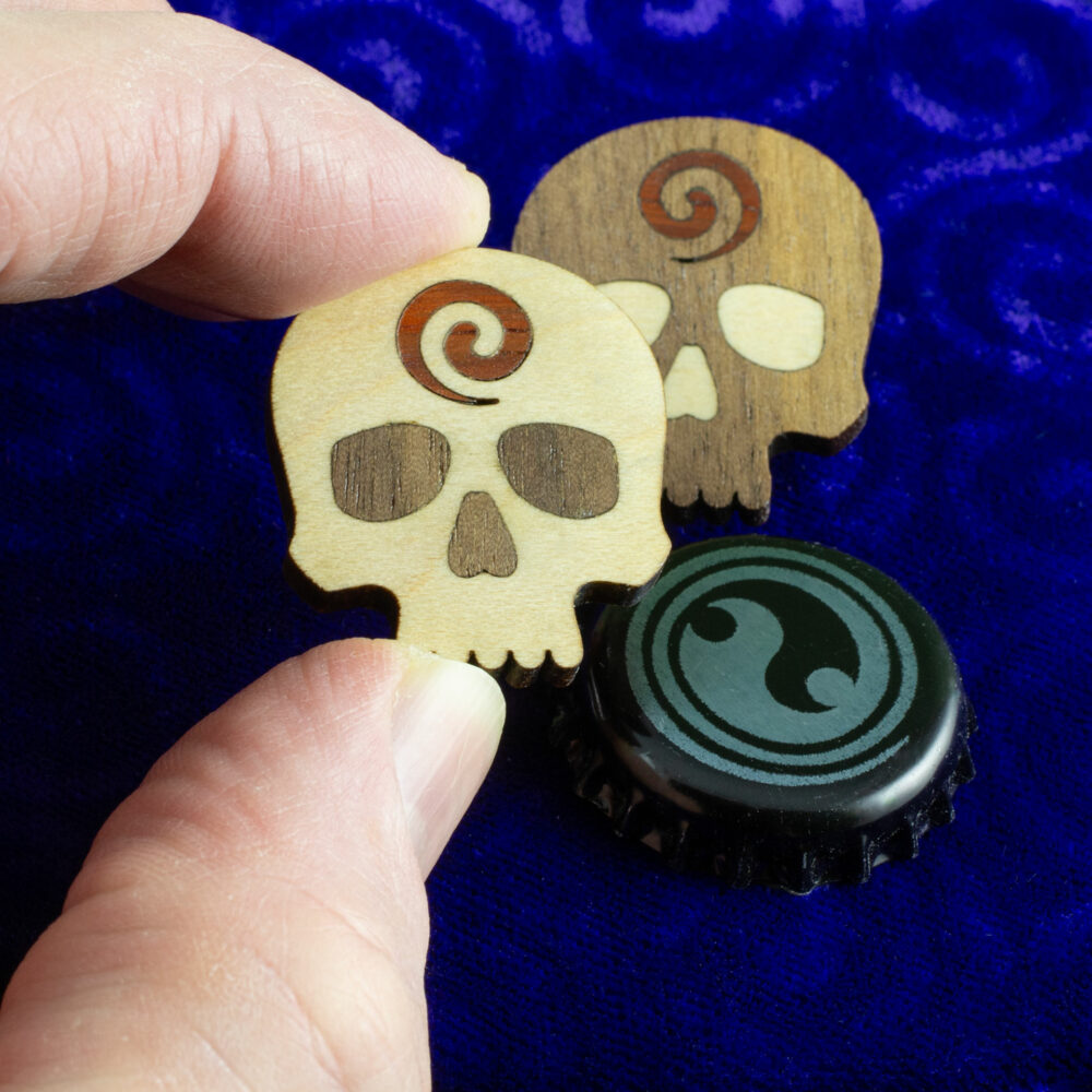 Godar Blot skull pins – hardwood inlay skull pins from white oak, walnut and, padauk. The white oak is dominant on one pin and the walnut is dominant on the other. A bottle cap slightly smaller than the pins is provided for scale and, the light pin is held between an index finger and thumb.