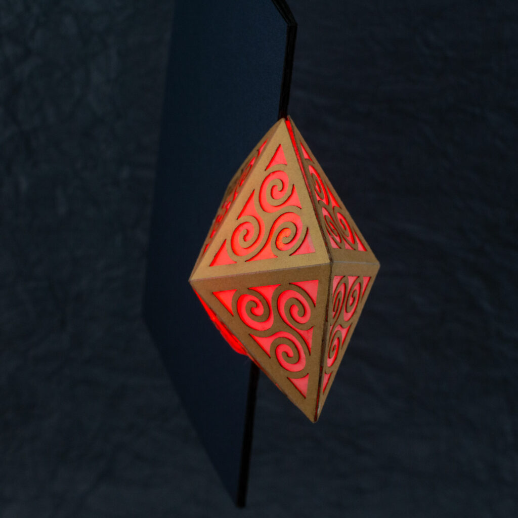 3/4 top-down view of the Spiral Crystal pop up card. Spiral Crystal pop up card fully open (360º). A yellow pentagonal lattice with triangular panels featuring Celtic triskelion designs is lit from within by a red light. The base card is a dark blue.