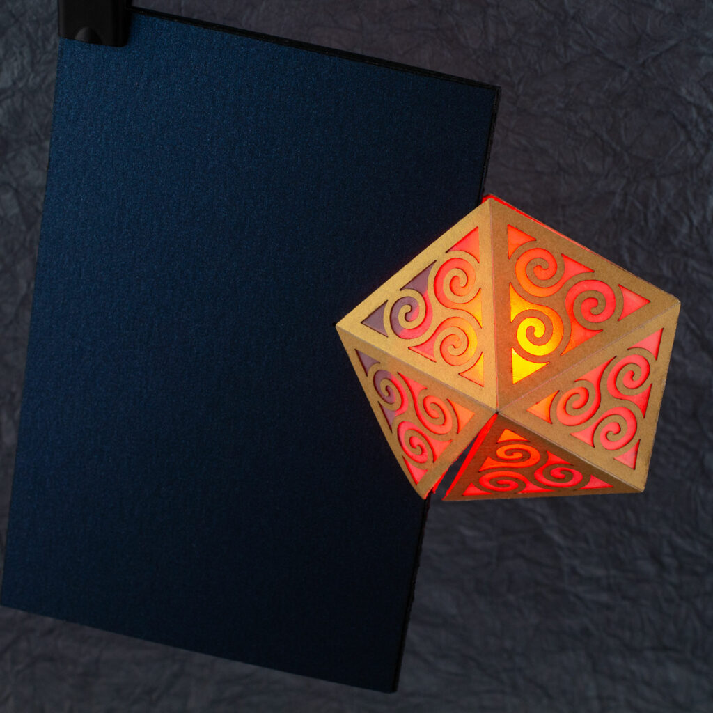 Spiral Crystal pop up card fully open (360º). A yellow pentagonal lattice with triangular panels featuring Celtic triskelion designs is lit from within by a red light. The base card is a dark blue.