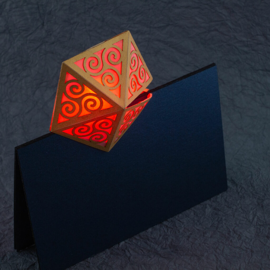 Spiral Crystal pop up card fully open (360º). A yellow pentagonal lattice with triangular panels featuring Celtic triskelion designs is lit from within by a red light. The base card is a dark blue.