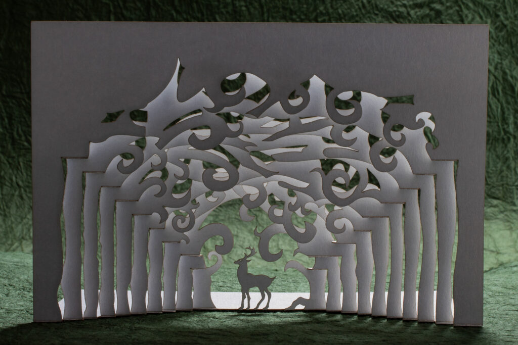 A pop up card made from cutting and folding a single sheet of stock depicts a stag standing framed by the convergence of rows of trees receding into the distance. The branches overhead form whirls and spirals. The background is a green textured paper (momigami) with the white card backlit against it.