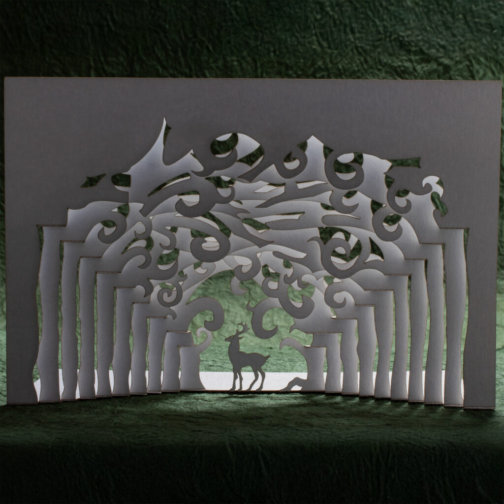 A pop up card made from cutting and folding a single sheet of stock depicts a stag standing framed by the convergence of rows of trees receding into the distance. The branches overhead form whirls and spirals. The background is a green textured paper (momigami) with the white card backlit against it.