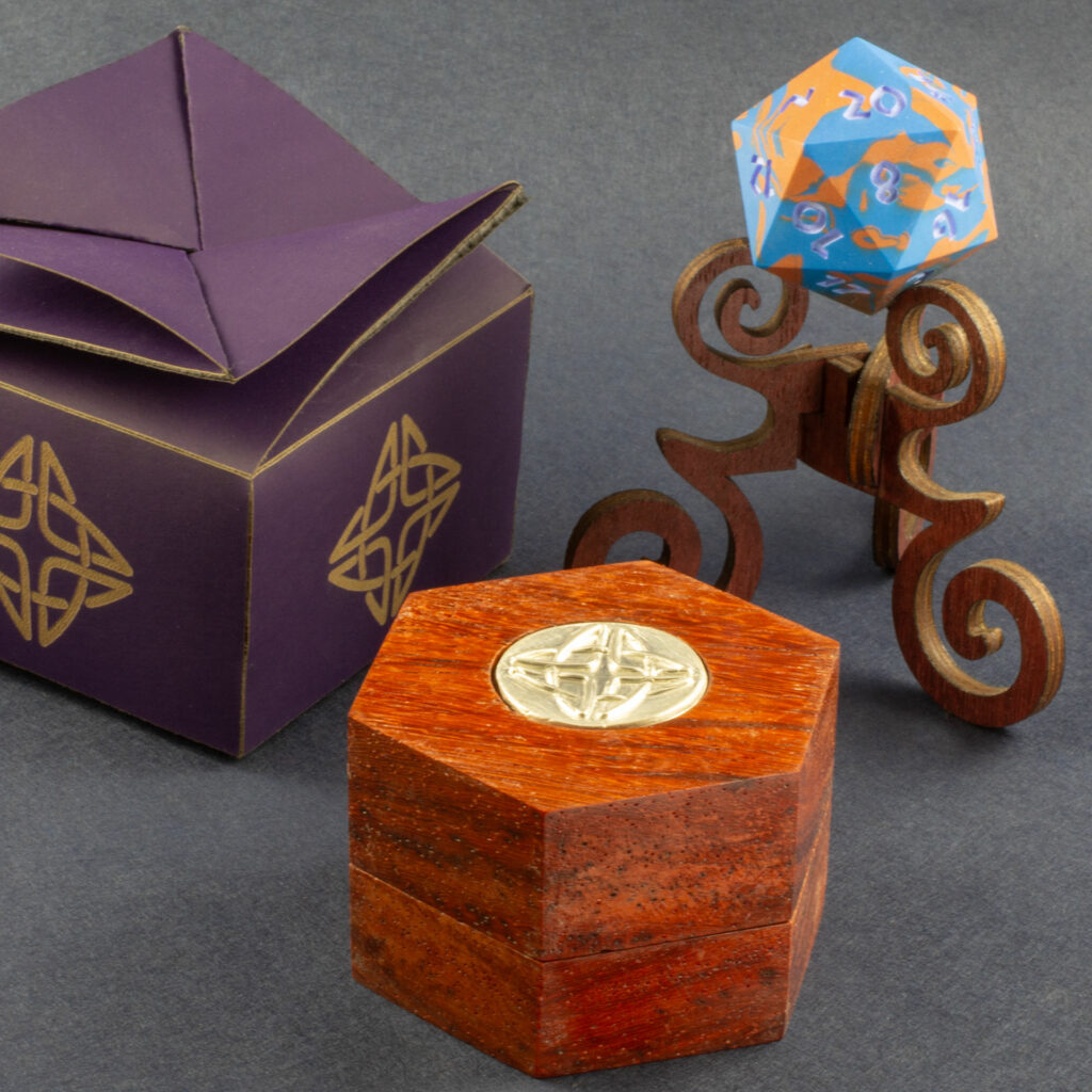 Padauk hardwood hexagonal box with a silver Celtic knotwork medallion in the lid sitting in front of a purpleheart wood stand bearing a blue and copper 42mm Jesmonite 20-sided die, and a purple twist-top box with matching knots engraved in the sides.