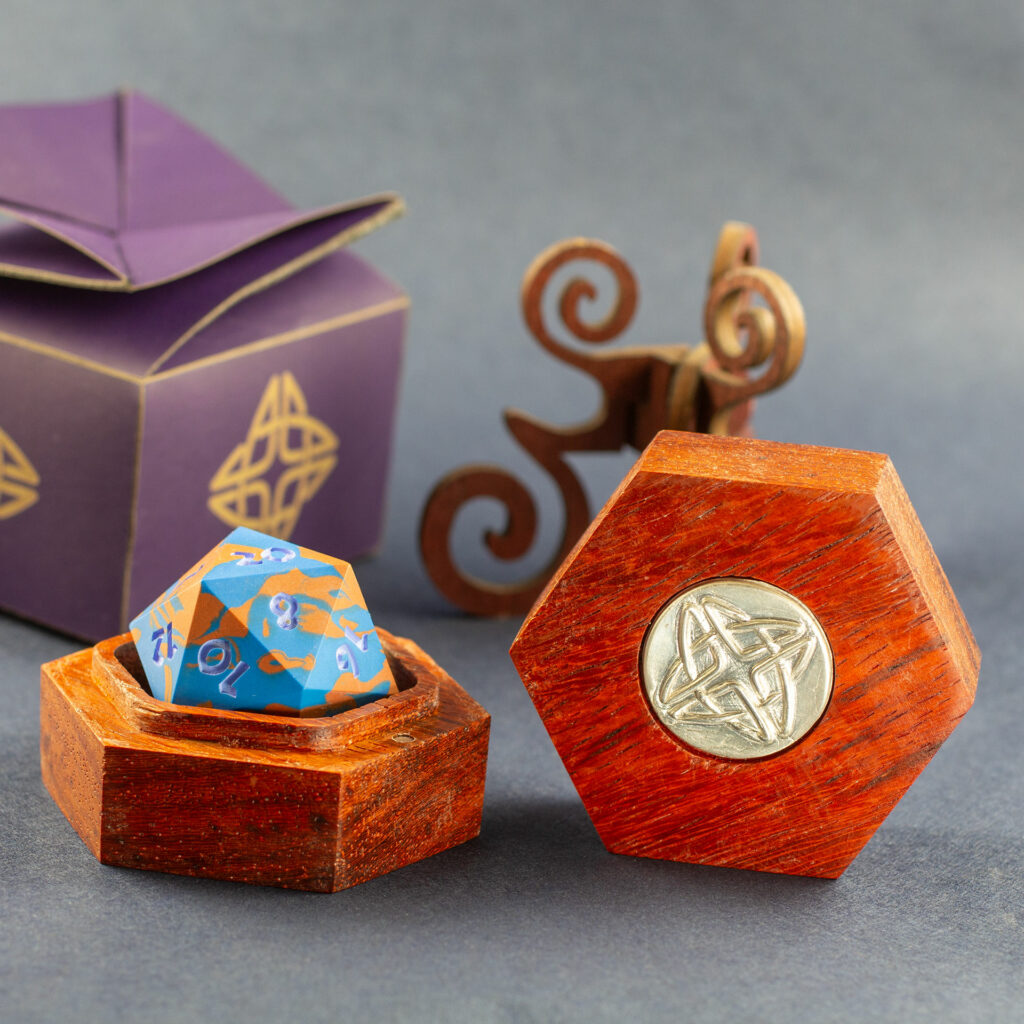 Padauk hardwood hexagonal box with a silver Celtic knotwork medallion in the lid, open with a blue and copper 42mm Jesmonite 20-sided die inside.