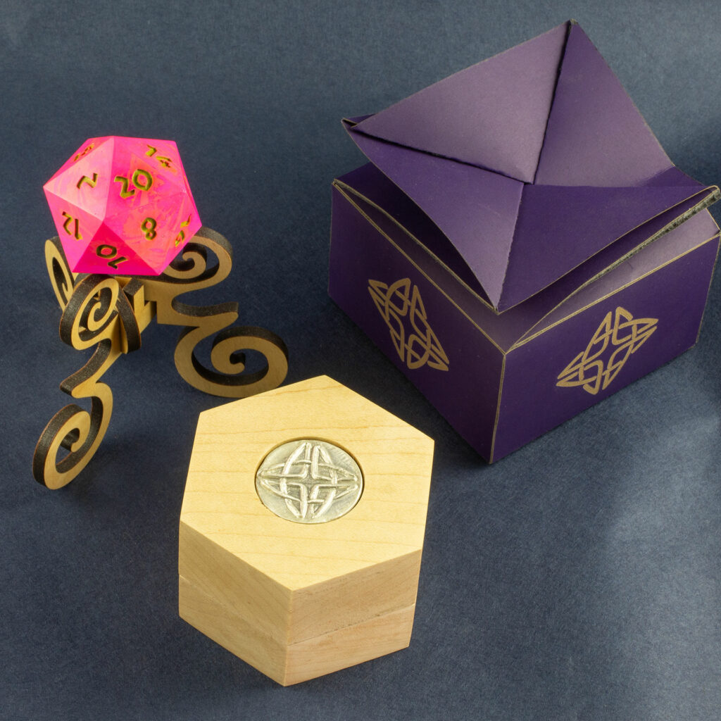 Oak Hex Chonk Box with a silver Celtic knotwork medallion in the lid, in front of an oak stand with a pink and orange 20-sided Jesmonite die, and a purple twist-top box.