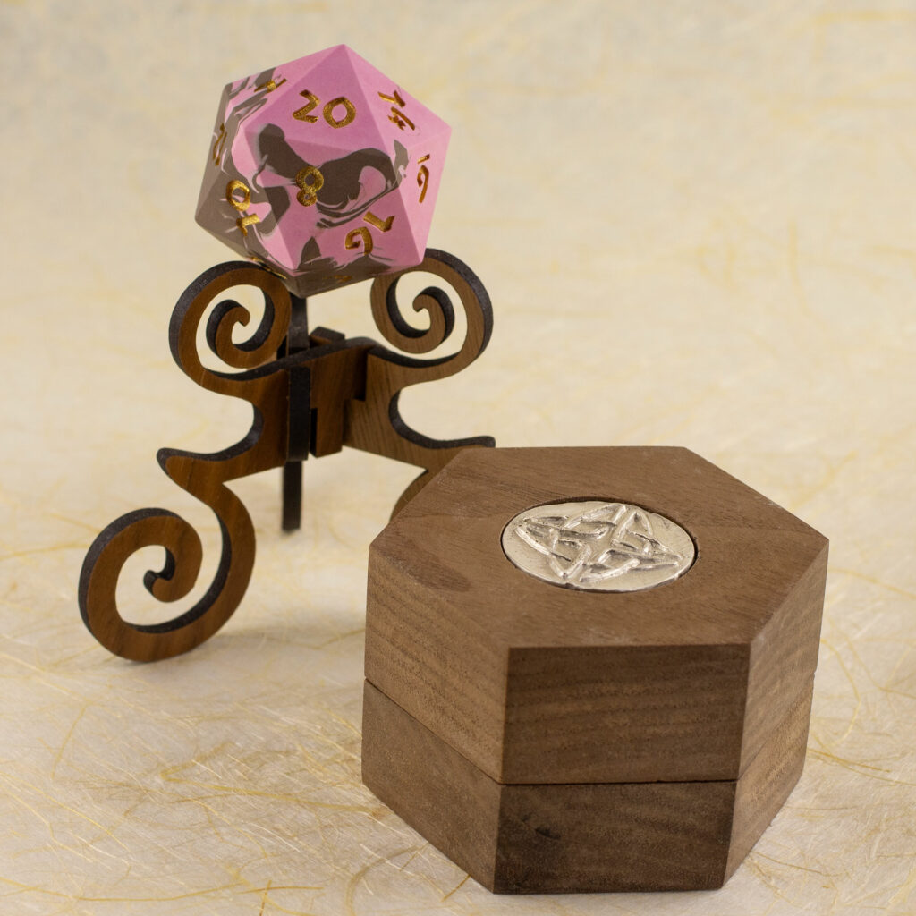 Walnut Hex Chonk Box with a silver Celtic knotwork medallion in the lid, in front of a walnut stand with a pink and brown 20-sided Jesmonite die.