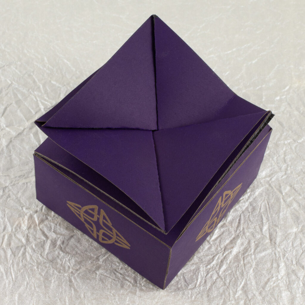 A small, squat, square, deep violet twist-top box with a Celtic knot engraved in a pale brown on each side (top down view).