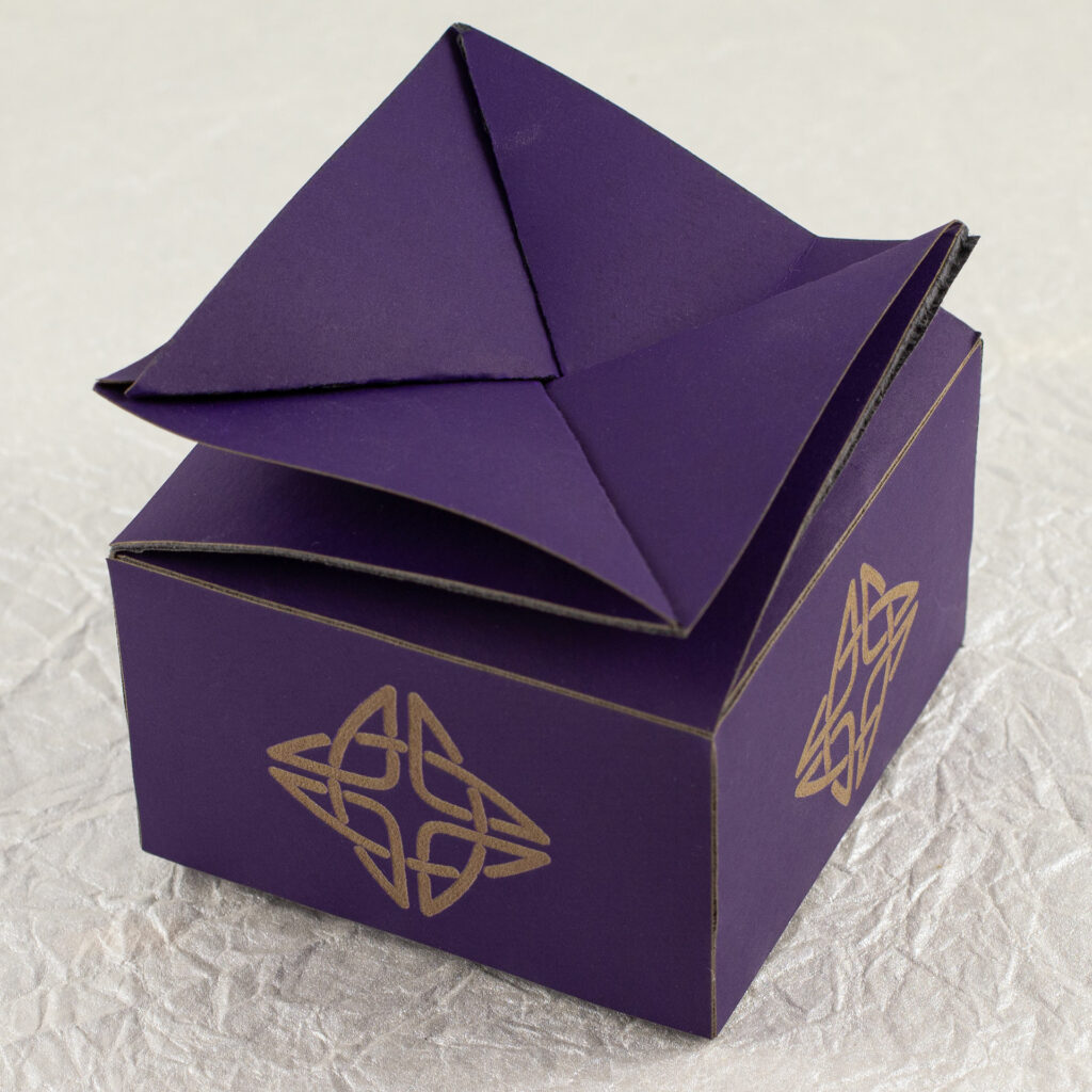 A small, squat, square, deep violet twist-top box with a Celtic knot engraved in a pale brown on each side (three-quarter top view).