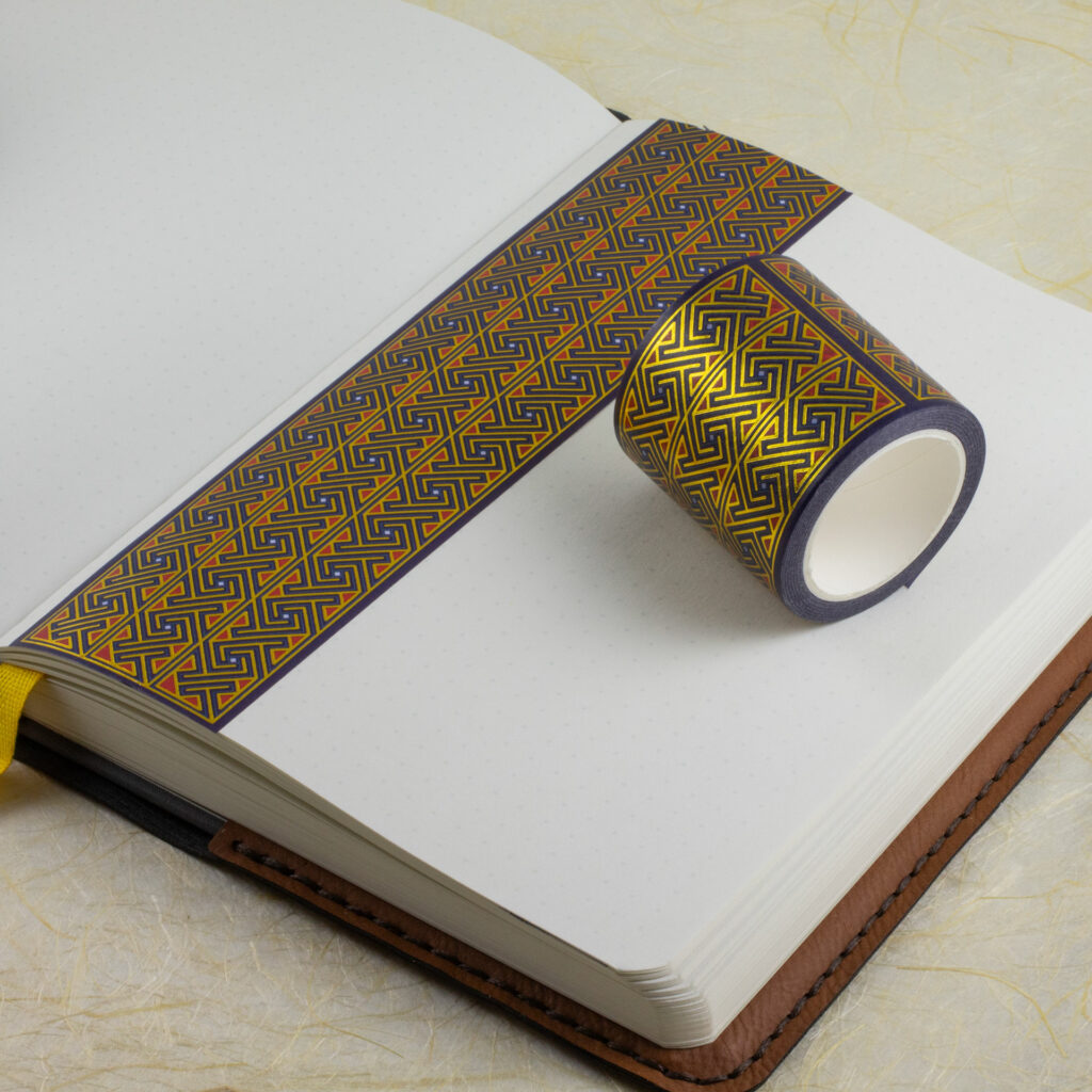 A roll of 45mm-wide Celtic key pattern washi tape sits on an open journal. A strip of the tape has been applied to a blank page along one edge.