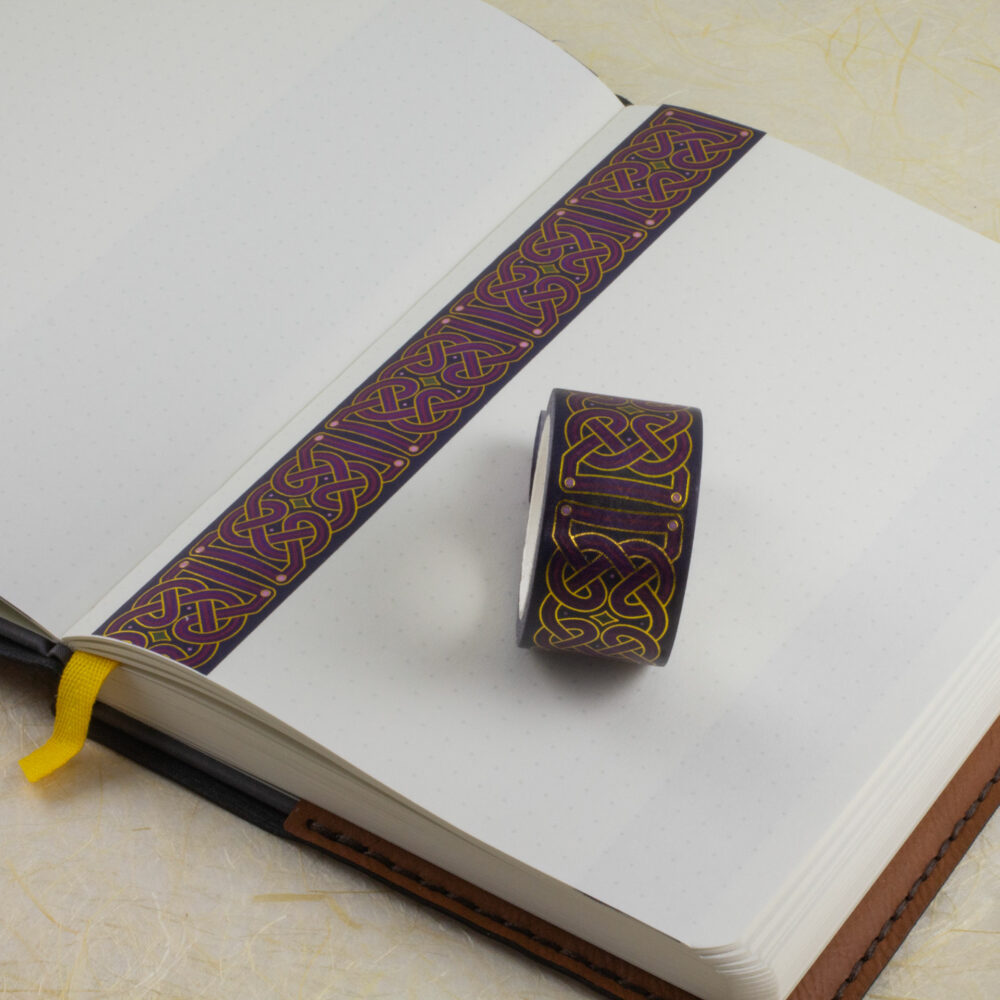 A roll of 25mm-wide Celtic knotwork washi tape sits on an open journal. A strip of the tape has been applied to a blank page along one edge.