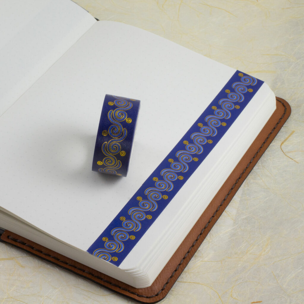 A roll of 20mm-wide washi tape with a Celtic spirals pattern sits on an open journal. A strip of the tape has been applied to a blank page along one edge.