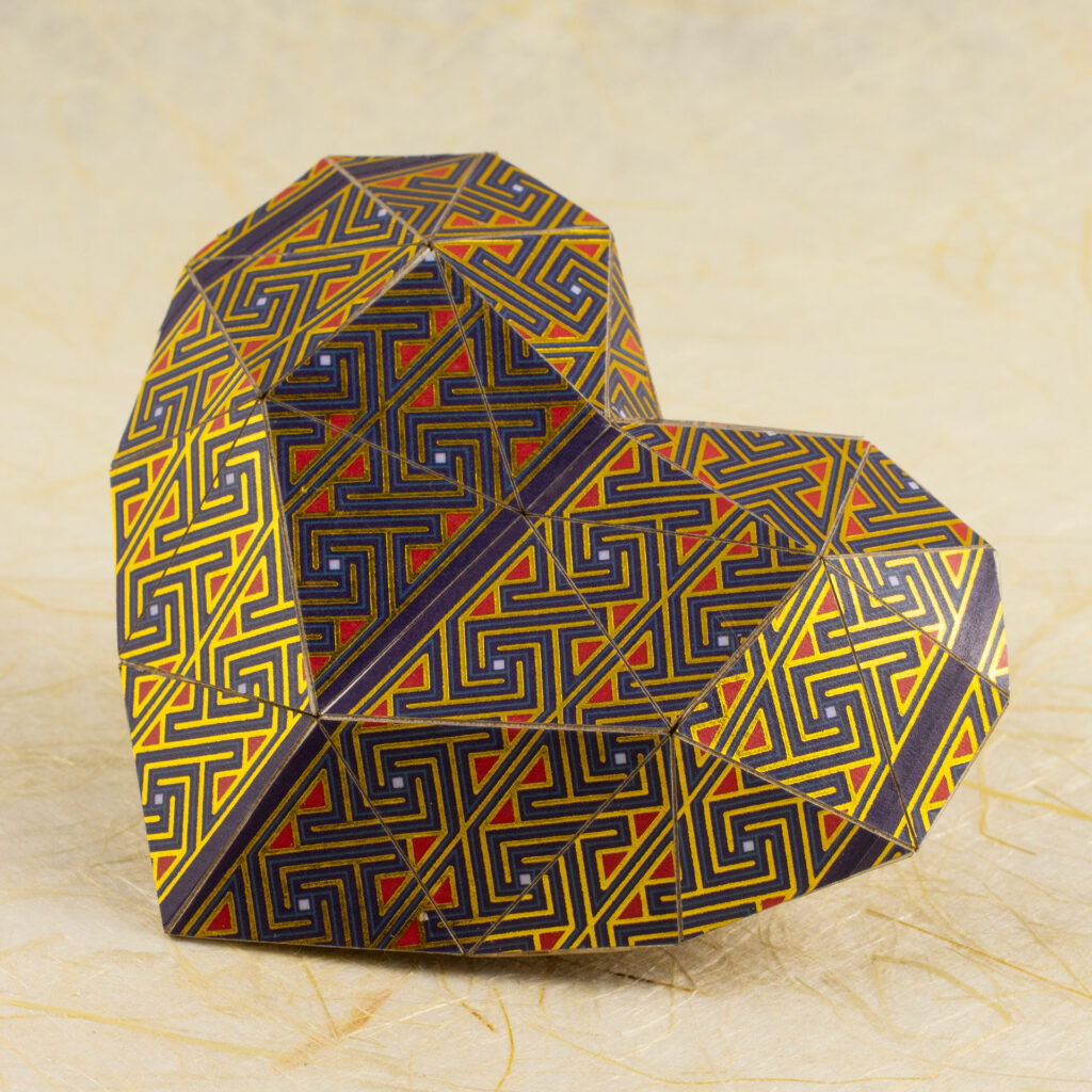 A pepakura (low poly) 3D paper heart covered in Celtic key/maze patterns in shades of purple, red, blue, and metallic gold rests on a cream-colored handmade paper background.