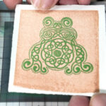 Frog celtic knotwork design printed in green on a copper square on white toothy paper being held up by the corners