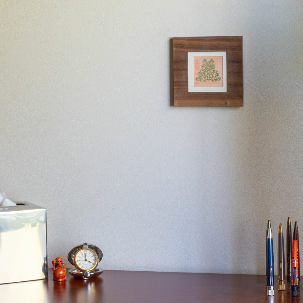 Frog Celtic knotwork print in a walnut frame hanging on the wall over a desk with a small clock, a netsuke of a skeleton playing a mokogyo, a silver tissue box, and a few Retro 51 pens standing point-up.