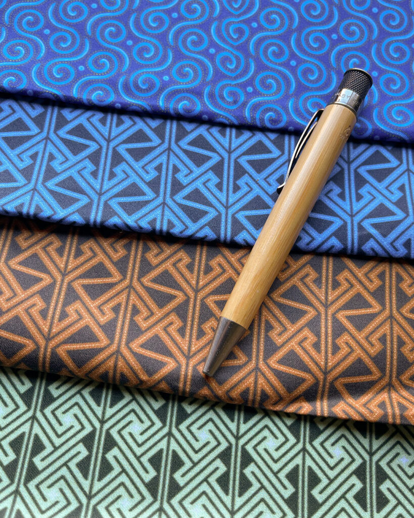 Four pieces of fabric arranged like shingles: a field of spirals in shades of blue, a Celtic key / maze pattern in an almost-black on blue, the same key / maze pattern on a warm brown, a different key /maze pattern (like squared spirals) in dark on a minty green. A bamboo body pen sits at an angle across the stack (for scale).