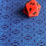 Fabric with an endless pattern of intricate Celtic knots in shades of blue with yellow stars on a darker blue field lying flat with a bright orange-red and bronze 20-sided die with black numbers.