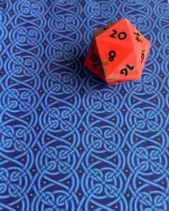 Fabric with an endless pattern of intricate Celtic knots in shades of blue with yellow stars on a darker blue field lying flat with a bright orange-red and bronze 20-sided die with black numbers.