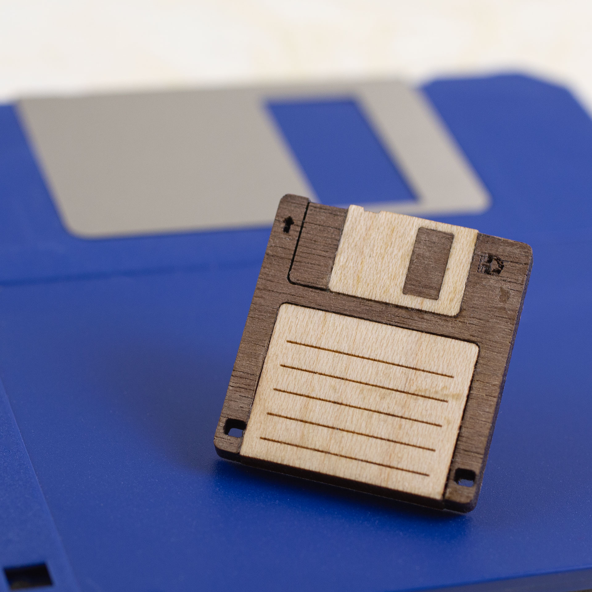 A small (24mm/1in square) pin that looks like a 3.5” floppy disk made from inlaid pieces of walnut and oak, sitting on a plain blue floppy disk.