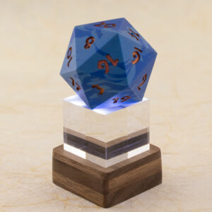 A one-inch clear acrylic cube sits in a small square layered walnut stand, which lights the cube and the 42 millimeter 20-sided die sitting on top of it from below. The die is a swirl of two shades of blue with metallic copper numbers. Shown against a defocused background of handmade paper with fiber inclusions. Side view.