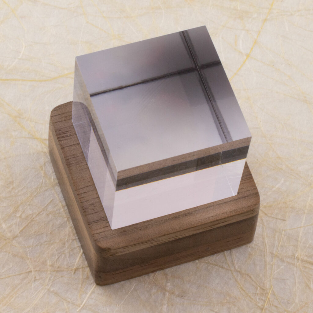 Three-quarter top-down view of a one-inch clear acrylic cube sitting in a small square layered walnut stand. The stand lights the cube from below, but the light is off in this image. Shown against a background of handmade paper with fiber inclusions.