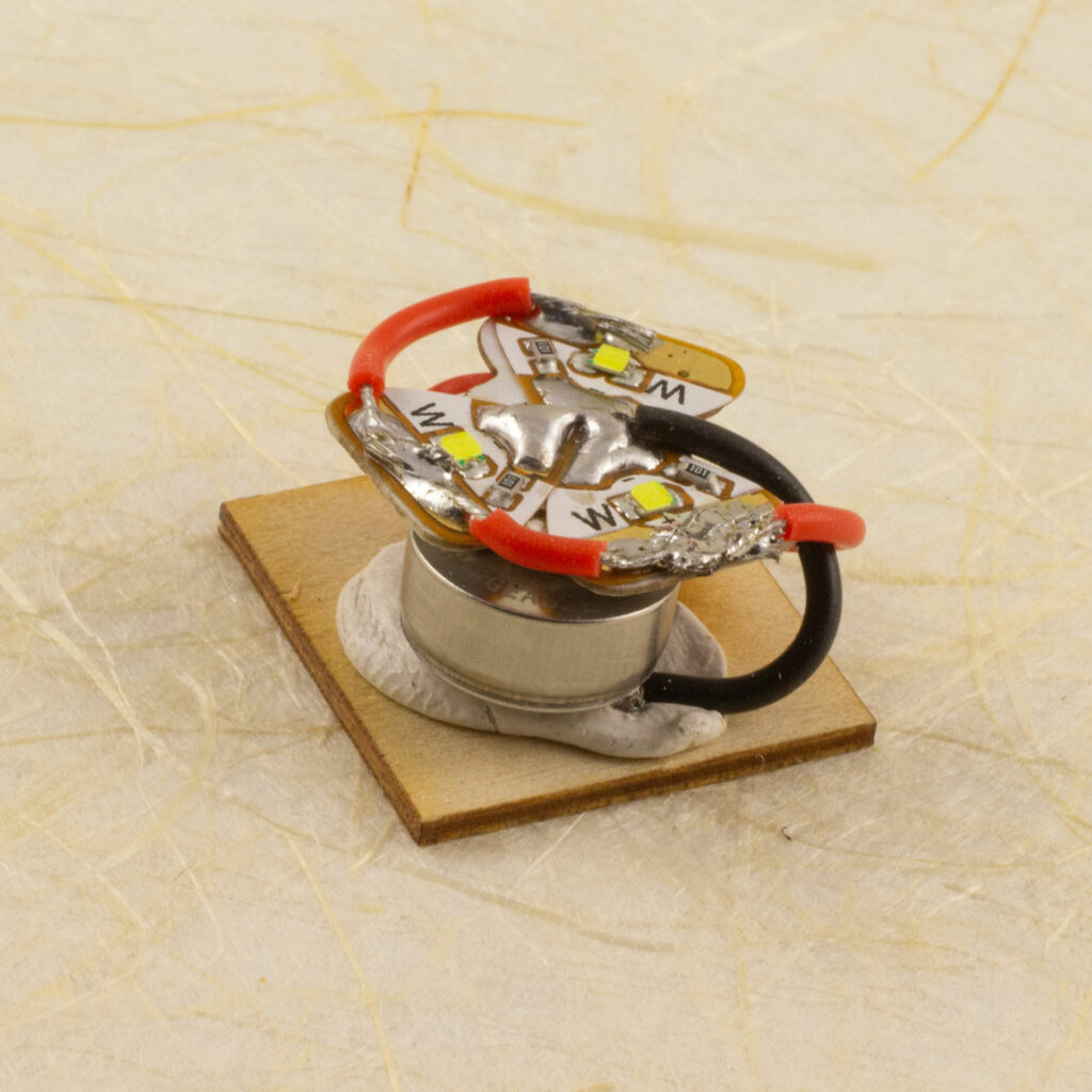 Electronics removed from the base of the small light-up stand showing three Chibitronics Circuit Stickers wired to a small rechargeable coin cell, stuck to the bottom panel with putty.