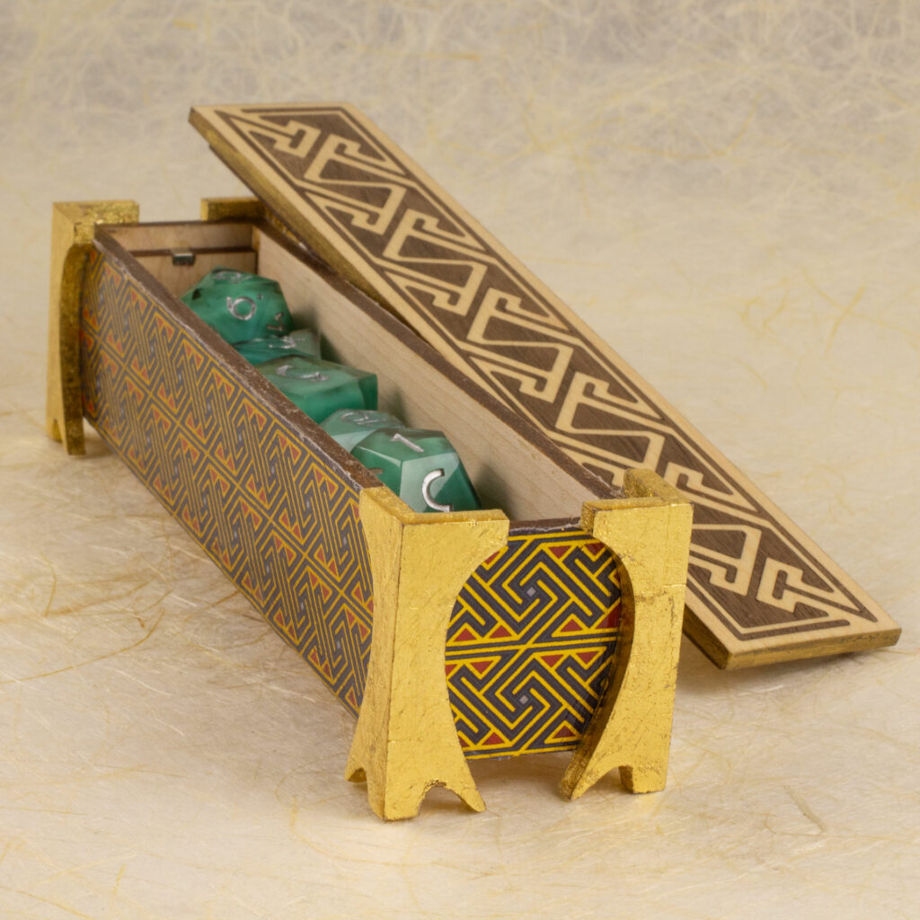 Dice vault (a long, thin box) with sides covered in a Celtic key pattern in blues, red, and metallic gold, attached gold-leafed neoclassical/nouveau-style legs, and a lid with a Celtic key pattern inlay in walnut and white oak against a background of off-wite handmade paper with fibrous inclusions. The box is open with the lid resting against it. Inside, a set of milky turquoise dice can be seen (three-quarter view).