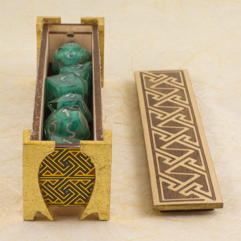 Dice vault (a long, thin box) with sides covered in a Celtic key pattern in blues, red, and metallic gold, attached gold-leafed neoclassical/nouveau-style legs, and a lid with a Celtic key pattern inlay in walnut and white oak against a background of off-wite handmade paper with fibrous inclusions. The box is open with the lid sitting next to it. Inside, a set of milky turquoise dice can be seen.