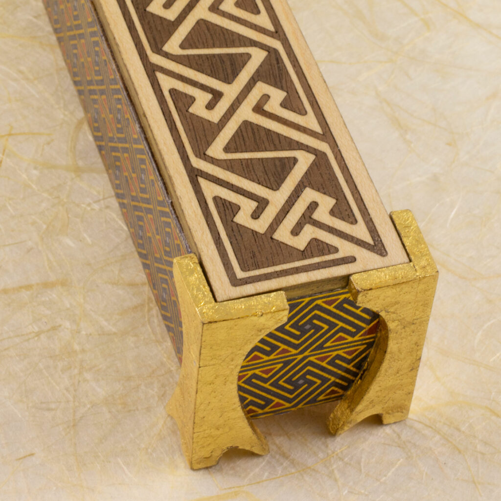 Dice vault (a long, thin box) with sides covered in a Celtic key pattern in blues, red, and metallic gold, attached gold-leafed neoclassical/nouveau-style legs, and a lid with a Celtic key pattern inlay in walnut and white oak against a background of off-wite handmade paper with fibrous inclusions (top view).