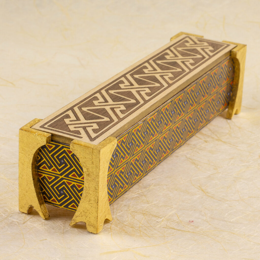 Dice vault (a long, thin box) with sides covered in a Celtic key pattern in blues, red, and metallic gold, attached gold-leafed neoclassical/nouveau-style legs, and a lid with a Celtic key pattern inlay in walnut and white oak against a background of off-wite handmade paper with fibrous inclusions (elevated three-quarter view looking).