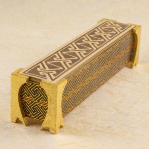 Dice vault (a long, thin box) with sides covered in a Celtic key pattern in blues, red, and metallic gold, attached gold-leafed neoclassical/nouveau-style legs, and a lid with a Celtic key pattern inlay in walnut and white oak against a background of off-wite handmade paper with fibrous inclusions (elevated three-quarter view looking).