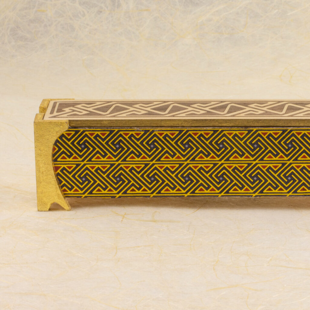 Dice vault (a long, thin box) with sides covered in a Celtic key pattern in blues, red, and metallic gold, attached gold-leafed neoclassical/nouveau-style legs, and a lid with a Celtic key pattern inlay in walnut and white oak against a background of off-wite handmade paper with fibrous inclusions (side view).