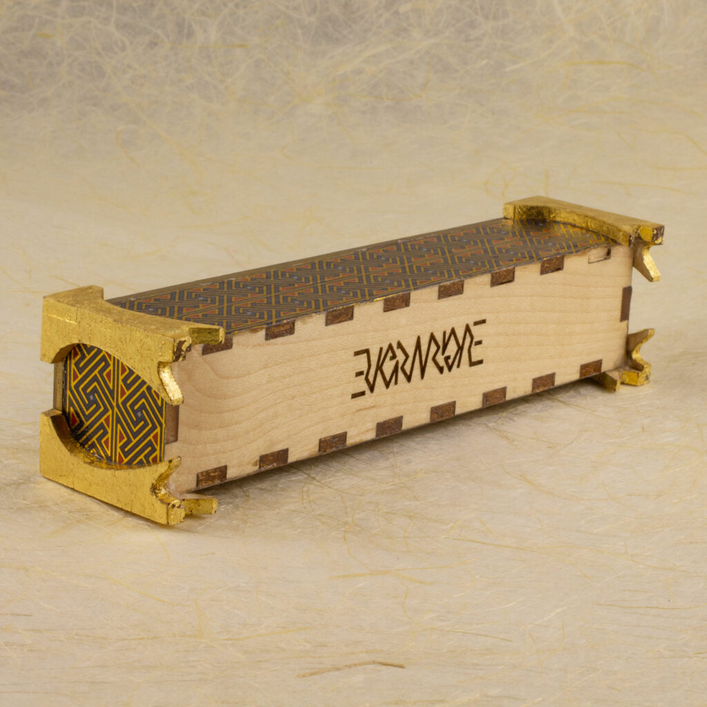 Three-quarter view of the Celtic key pattern dice vault on its side showing the bottom, which has been engraved with the Evermore ambigram, on a surface and background of handmade off-white paper.