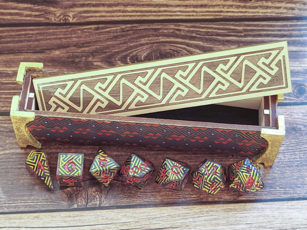 Dice vault (a long, thin box) with sides covered in a Celtic key pattern in blues, red, and metallic gold, attached gold-leafed neoclassical/nouveau-style legs, and a lid with a Celtic key pattern inlay in walnut and white oak on a faux wood surface. The box is open with the lid sitting at an angle inside the box. In front of the box are a set of dice made with the same washi tape used on the box exterior (front top-down view).