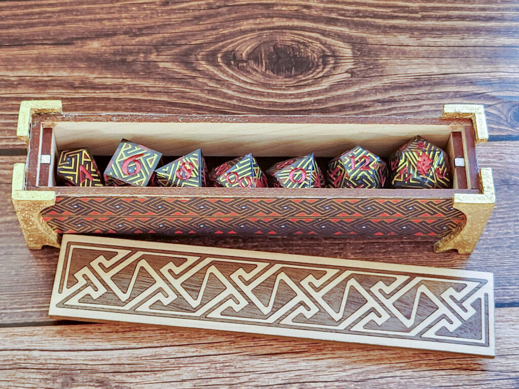 Dice vault (a long, thin box) with sides covered in a Celtic key pattern in blues, red, and metallic gold, attached gold-leafed neoclassical/nouveau-style legs, and a lid with a Celtic key pattern inlay in walnut and white oak on a faux wood surface. The box is open with the lid sitting in front of it. Inside, a set of dice made with the same washi tape used on the box exterior can be seen (front top-down view).