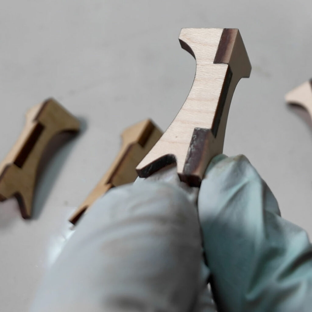 A gloved hand holds up a wooden box leg showing off the design change making the foot have less of a point.